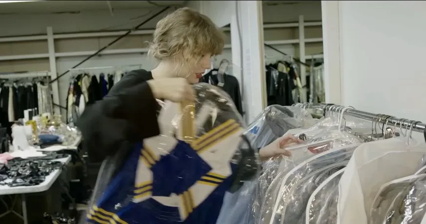 Taylor Swift lifting a blue, gold and white cheerleading uniform from the clothing rack