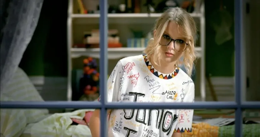 Taylor Swift in black-rimmed glasses and a handwritten Junior Jewels T-shirt behind a window frame