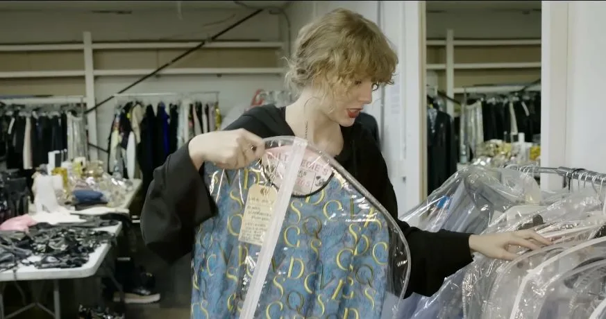 Taylor Swift displaying a blue bodysuit decorated with gold letters inside a clear garment bag