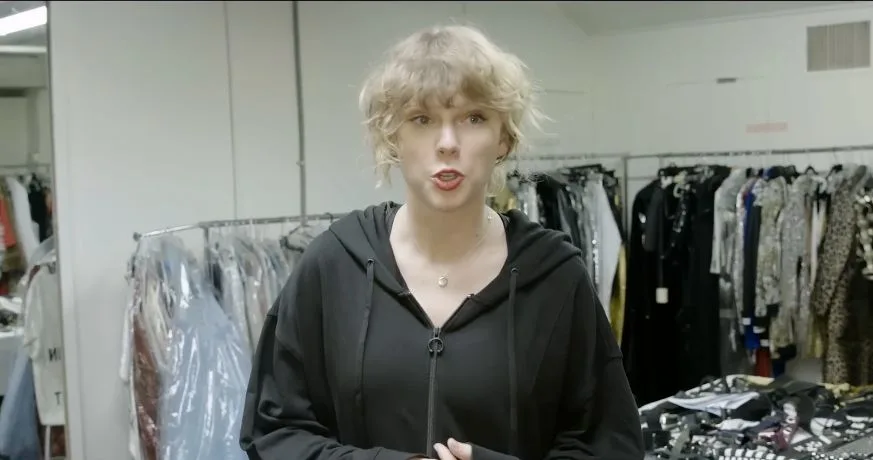 Taylor Swift surrounded by garment racks and a table of accessories in the wardrobe room